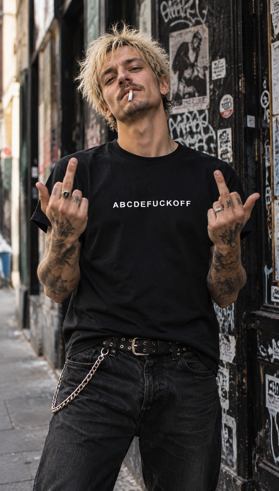 Tattooed man with bleached messy hair wearing a black ABCDEFUCKOFF T-shirt, raising both middle fingers against a graffiti-covered urban doorway.