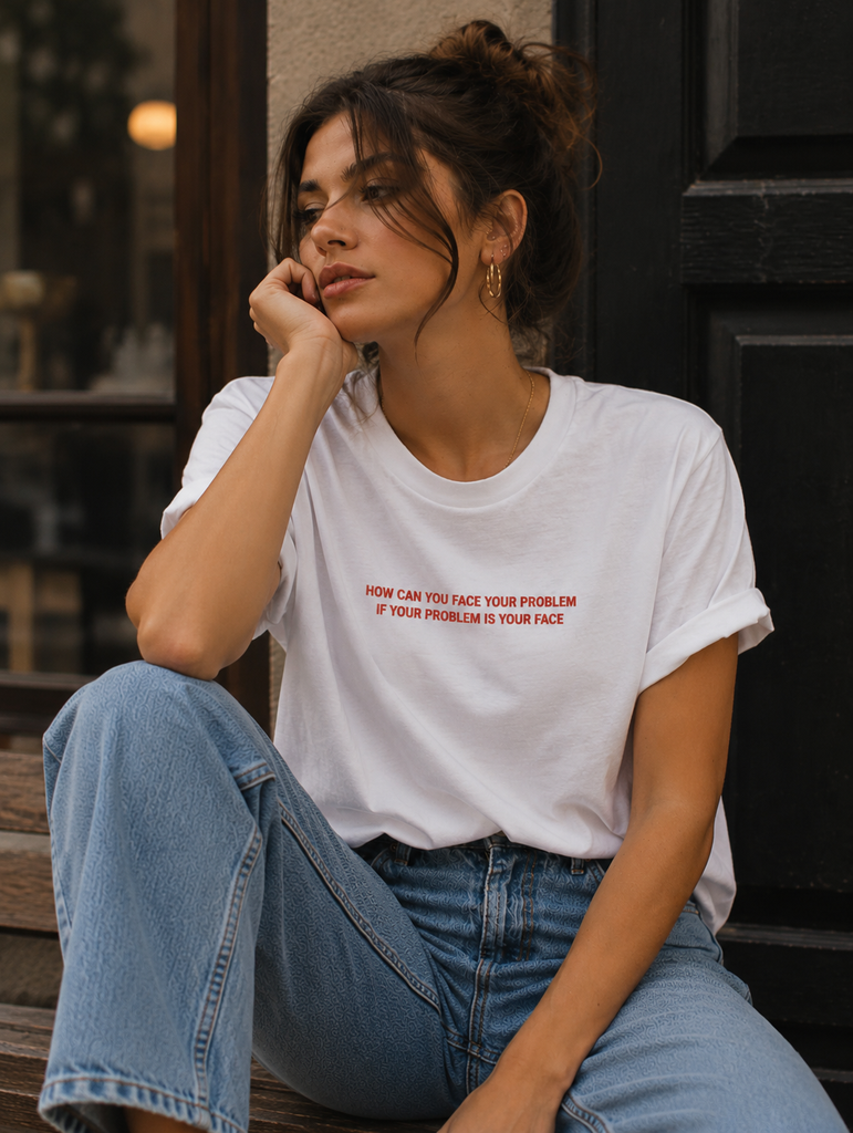 A woman sitting on wooden steps in front of a dark doorway, wearing a white graphic t-shirt with red text that reads 'HOW CAN YOU FACE YOUR PROBLEM IF YOUR PROBLEM IS YOUR FACE', paired with light blue jeans.