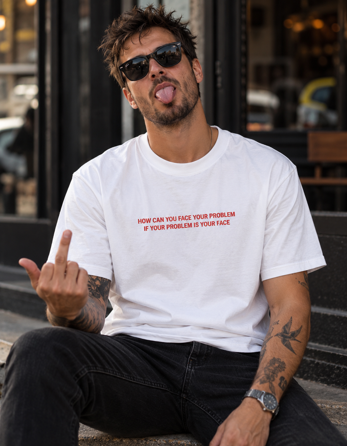 A man wearing a white graphic t-shirt with red text that reads 'HOW CAN YOU FACE YOUR PROBLEM IF YOUR PROBLEM IS YOUR FACE.' He is sitting outdoors on stone steps, wearing black sunglasses and black jeans.