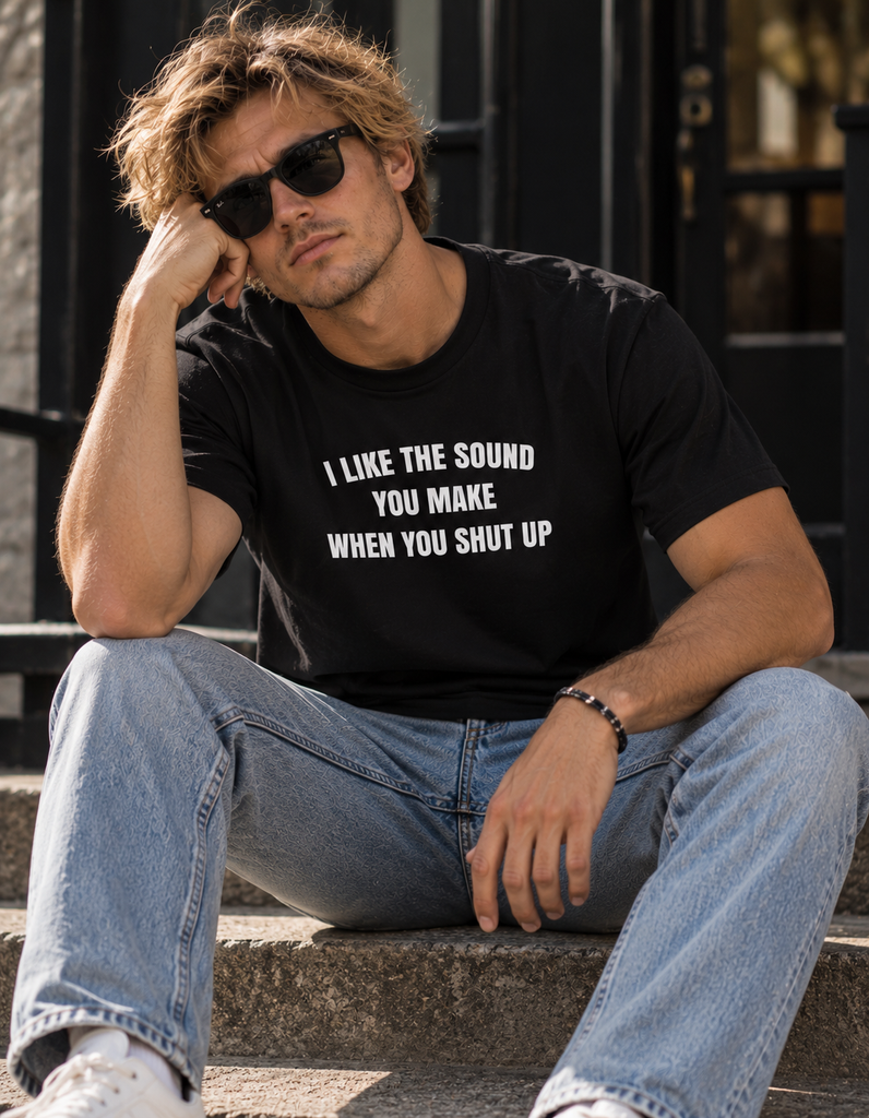 A man wearing a black graphic t-shirt with white text that reads 'I LIKE THE SOUND YOU MAKE WHEN YOU SHUT UP', paired with light blue jeans and sunglasses, sitting on outdoor stone steps.