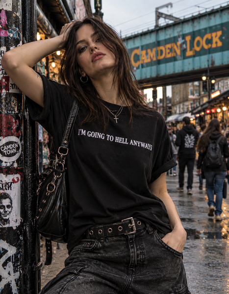 A woman posing on a city street wearing a black graphic t-shirt with the white text 'I'M GOING TO HELL ANYWAY'. She is leaning against a graffiti-covered pillar, with a black shoulder bag and dark jeans in a wet, urban setting under a blue Cambridge Lock bridge.