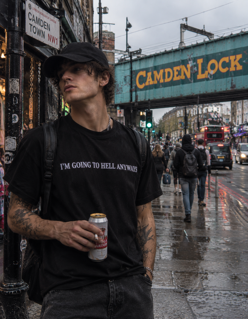 A man wearing a black graphic t-shirt with the white text 'I'M GOING TO HELL ANYWAYS' and a black cap, standing on a wet city street in London under a green Cambridge Lock bridge.