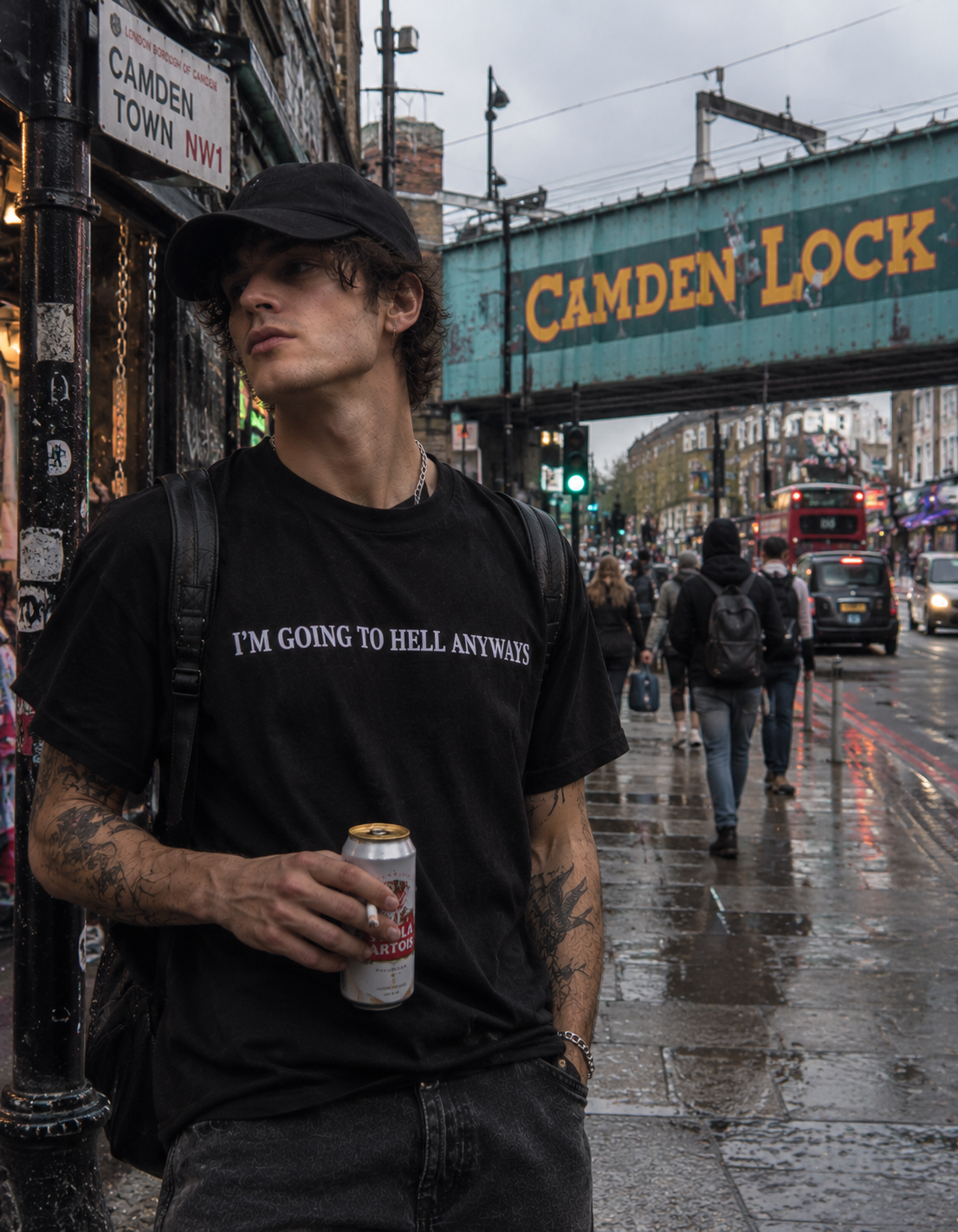 A man wearing a black graphic t-shirt with the white text 'I'M GOING TO HELL ANYWAYS' and a black cap, standing on a wet city street in London under a green Cambridge Lock bridge.