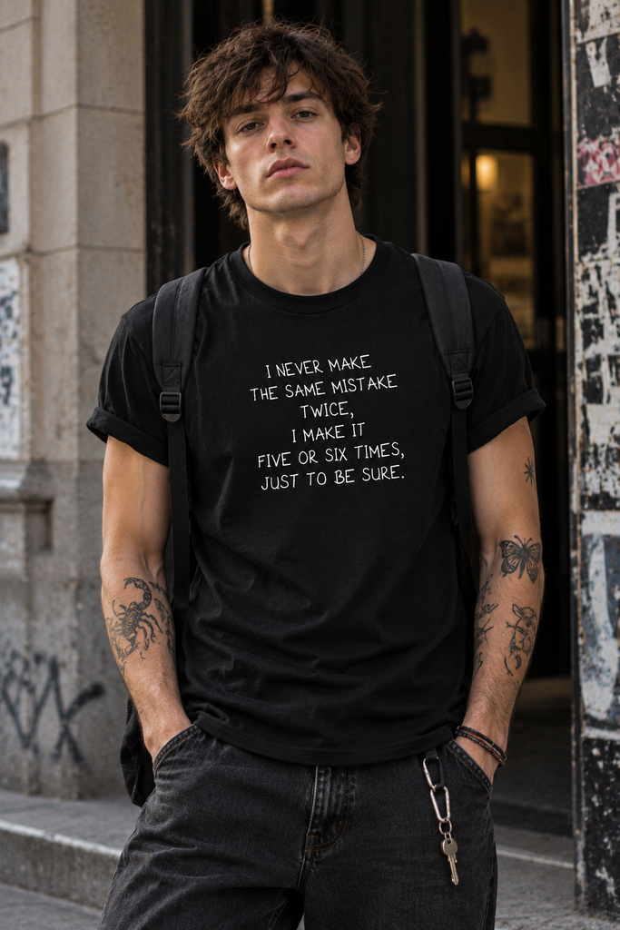 A man wearing a black graphic t-shirt with white text that reads 'I NEVER MAKE THE SAME MISTAKE TWICE, I MAKE IT FIVE OR SIX TIMES, JUST TO BE SURE.' He is standing outdoors in front of a building wall with posters.