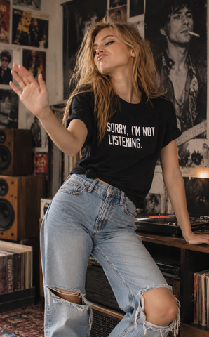 A woman in a music room wearing a black graphic t-shirt that reads 'SORRY, I'M NOT LISTENING.' and light-wash ripped jeans.