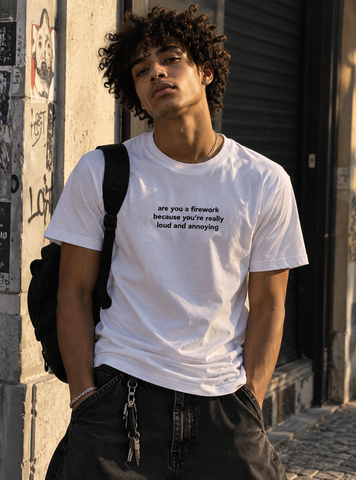 A man wearing a white graphic t-shirt with black text that reads 'are you a fireworks because you're really loud and annoying', paired with black jeans and a black backpack, standing in an outdoor urban alleyway.