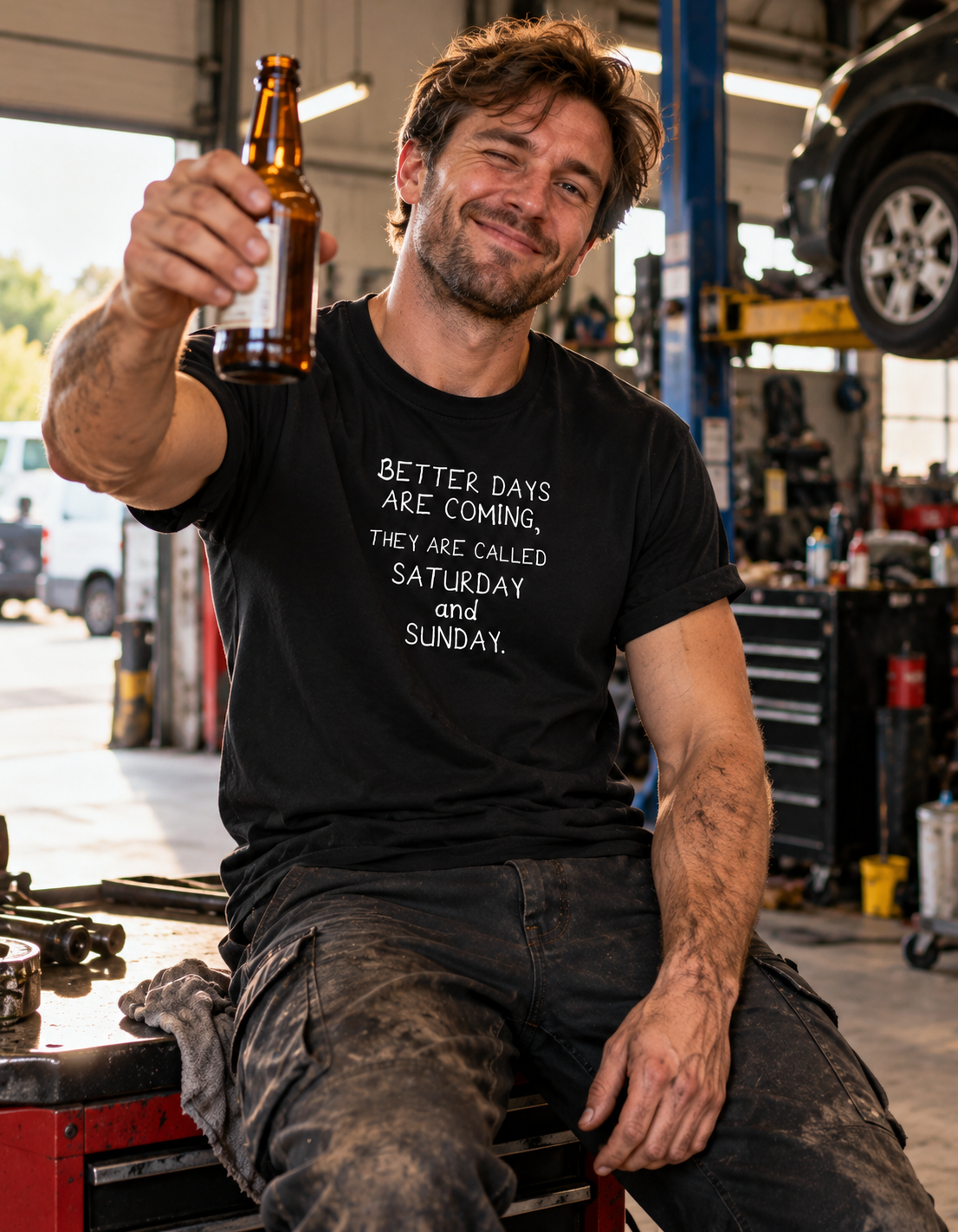 A man in a garage wearing a black graphic t-shirt that reads 'BETTER DAYS ARE COMING, THEY ARE CALLED SATURDAY and SUNDAY.' He is holding up a brown glass beer bottle.