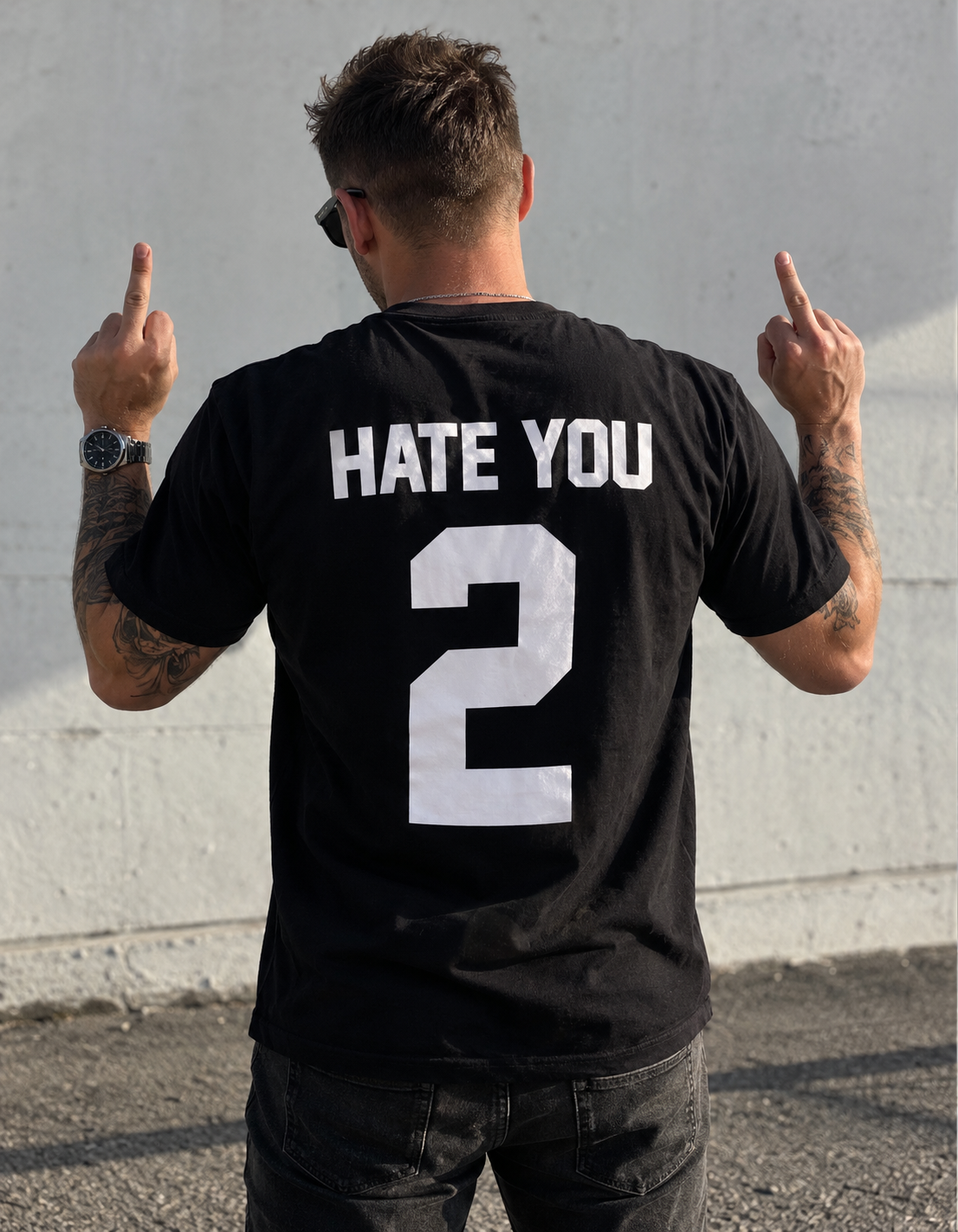Tattooed man wearing a black Hate You 2 rebellious slogan T-shirt, showing both middle fingers with the white back print clearly visible