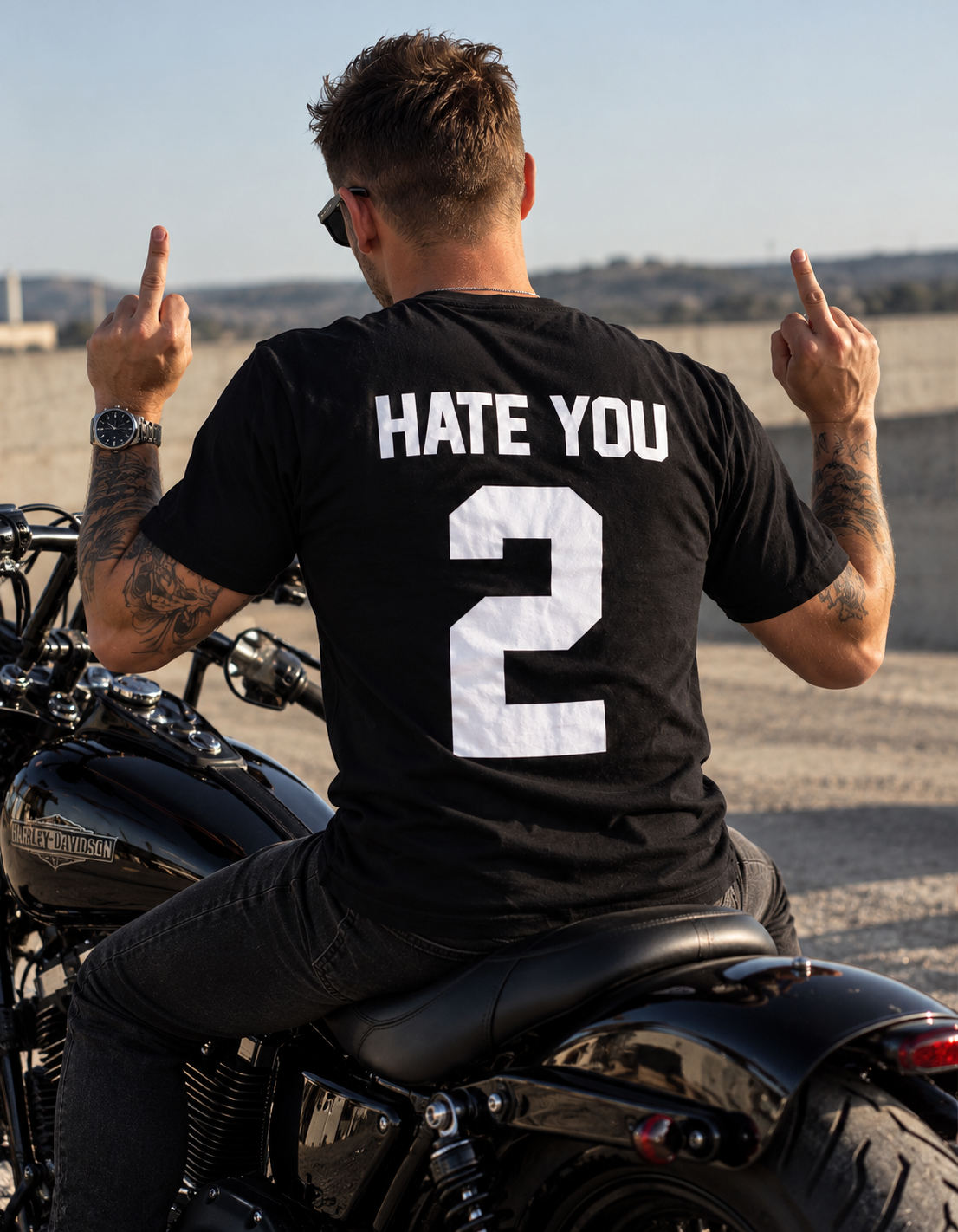A man sitting on a black motorcycle, viewed from behind, wearing a black t-shirt with 'HATE YOU' and the number '2' printed in white on the back. He is making middle-finger gestures with both hands in an outdoor setting.