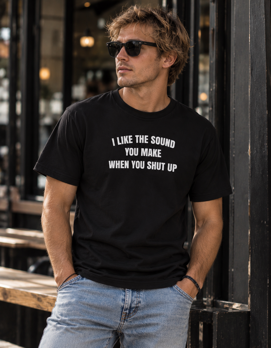 A man wearing a black graphic t-shirt with the white text 'I LIKE THE SOUND YOU MAKE WHEN YOU SHUT UP' and light blue jeans, standing in front of a dark storefront.