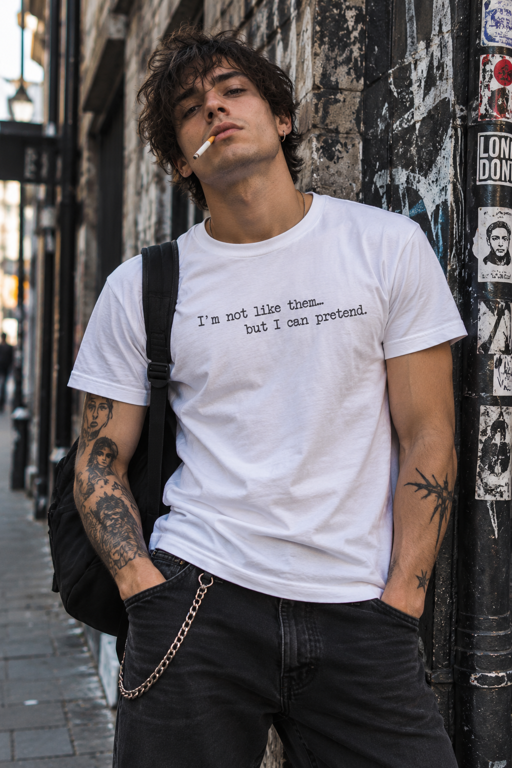 A man with tattoos leans against a city wall wearing a white graphic t-shirt that reads 'I'm not like them.