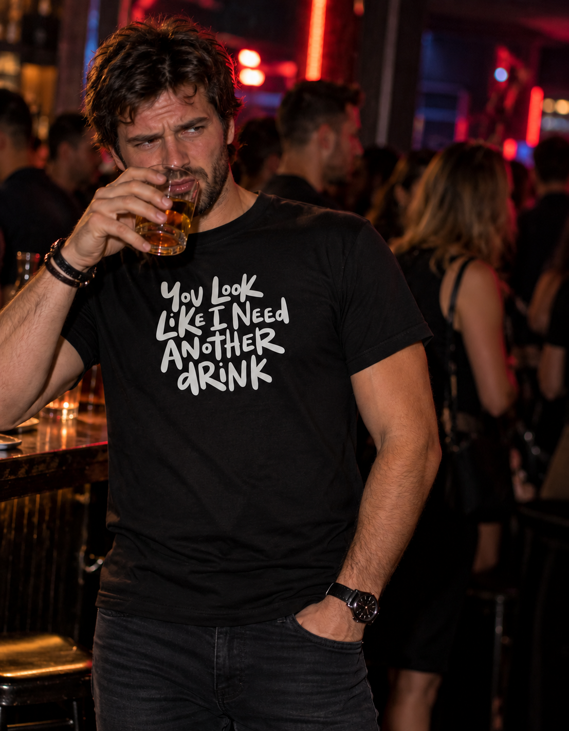 A man in a dimly lit bar wearing a black graphic t-shirt with the white text 'YOU LOOK LIKE I NEED ANOTHER DRINK'.